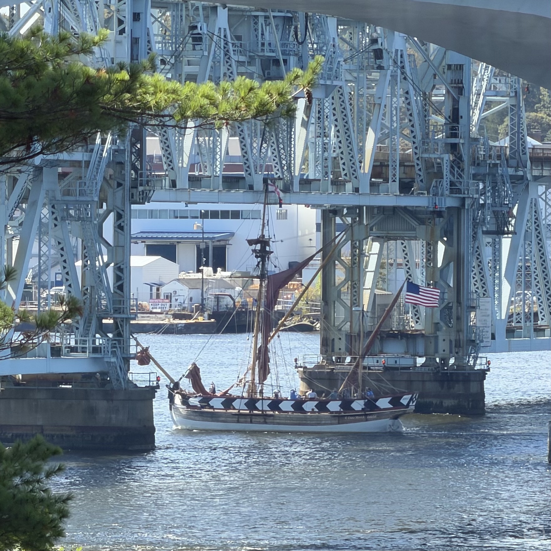 The wooden sailing vessel Virginia, with a red sail and an American flag flying, passing between the steel lift-span towers of the Carlton Bridge in Bath, Maine